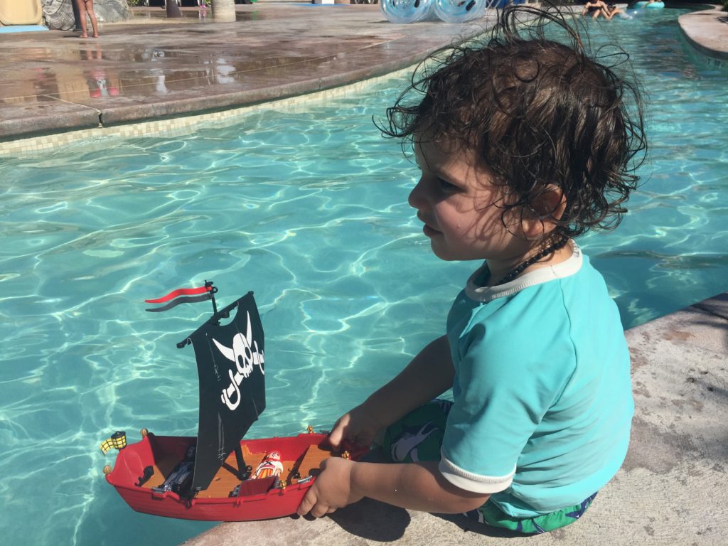 toddler boy wearing teal coloured rash guard sits on the edge of the lazy river with a toy pirate ship in his hands at Beaches Turks and Caicos Resort